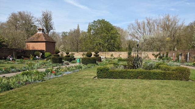 A view of a walled garden on a sunny day; several short-cut lawns with flowerbeds around the edges, and hedges and topiaries. It's all surrounded by an old-looking red brick wall. Behind the wall are trees and a blue sky with thin clouds. Forming part of the wall is a square brick-walled structure with pitched roof.