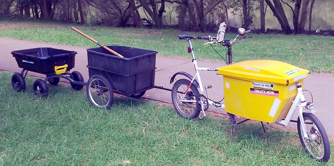 A long john style cargo bike towing a two wheel trailer that has a big black plastic bin, then towing behind that trailer a four wheel garden cart. It's very long. On the grass next to a concrete path with mangroves and a river in the background