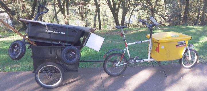 a long john style cargo bike towing a two wheel trailer, with a four wheel garden cart tied to the top. On a concrete path then in the background grass then mangroves and a river