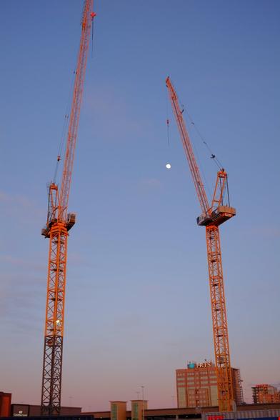 Two cranes in front of the moon appearing in the still blue sky. The crane on the right is positioned so that it tries to grab the moon.
