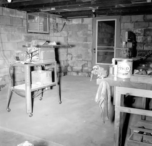 A basement with a concrete floor and cinder block walls. A workbench can be seen with a vise, some tools, and a can of Crisco. Two other large tools (perhaps a table saw and a drill or jigsaw). There is a window high on one wall and a door on the far wall with stairs beyond it.