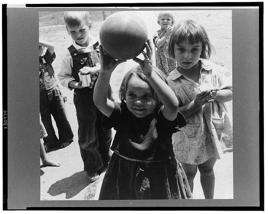 The image is a black and white photograph depicting several young children outdoors, engaging in play. One child in the foreground holds a large ball above her head with both hands, smiling at the camera. She wears a dark dress with ruffled sleeves and has light-colored hair pulled back from her face.

Behind her, there are three other children of varying ages; one to her left is wearing overalls holding a container, another beside him appears focused on something out of frame, while a girl in floral clothing stands slightly behind the central child. The setting seems informal with open space and what looks like unpaved ground beneath their feet.

The photograph has an air of candidness and simplicity, capturing a moment from everyday life without any apparent staging or formal composition. It is labeled as part of historical records related to migrant agricultural workers in California, specifically within Tulare County, possibly taken at a camp associated with the Farm Security Administration (FSA) which aimed to document rural American communities during the Great Depression era.

The image's source and additional information can be found online through various archives or collections dedicated to Dorothea Lange's work.