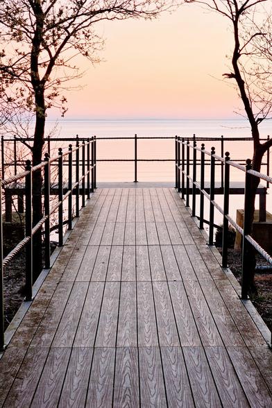 A wooden boardwalk with black railings leads towards a calm body of water under a soft pink and orange sky at sunset. Bare trees frame the walkway.
