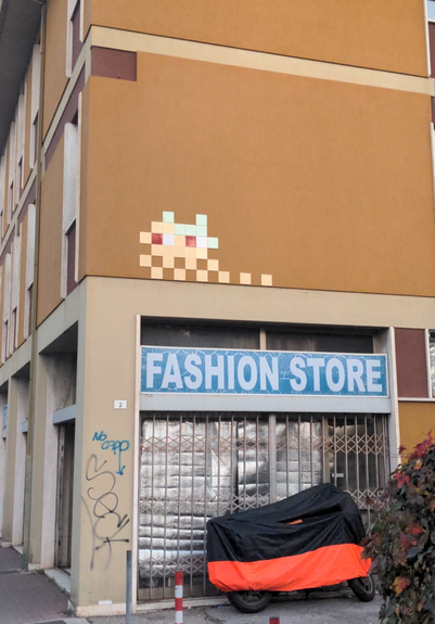 À un coin de rue, sur un mur orange ocre au-dessus de la boutique "fashion store", un grand invader dont les pixels sont représentés par des carreaux blancs, jaunes et saumon, les yeux rouges.