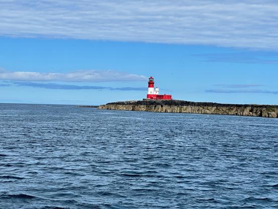 This image depicts a coastal scene with a lighthouse situated on a rocky outcrop. The lighthouse is painted red and white and stands prominently against the backdrop of a partly cloudy sky. The sea is calm, with gentle waves stretching across the foreground, while the rocky cliff extends along the horizon, leading to the lighthouse.