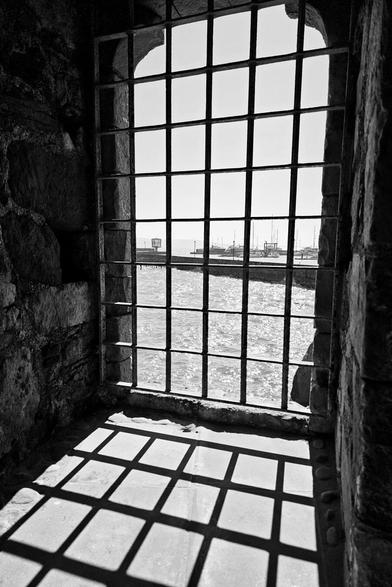 A black and white photograph shows a view of a harbor with boats and water seen through a barred window set in a stone wall. The sunlight casts a grid-like shadow on the floor in front of the window.