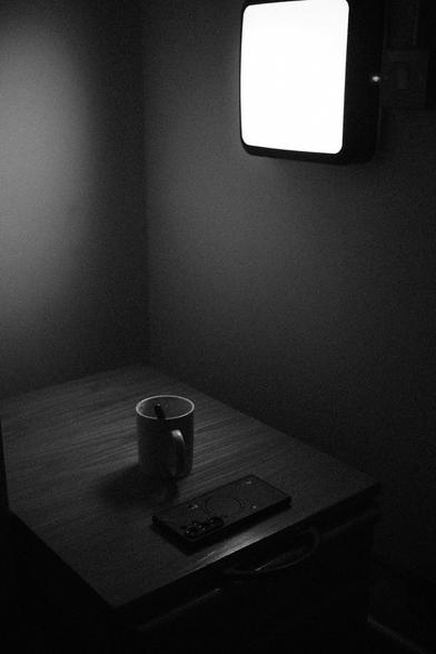 A monochromatic photograph depicts a cup of coffee illuminated by a wall-mounted lamp.