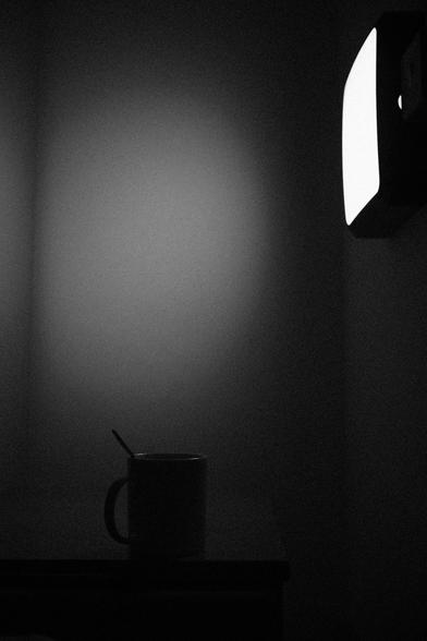 A monochromatic photograph depicts a cup of coffee illuminated by a wall-mounted lamp.