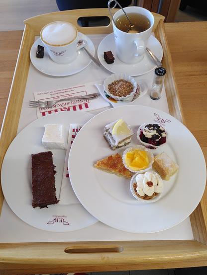 Auf einem großen Tablett stehen zwei Teller mit Kuchen: links ein klassischer Brownie und ein Marshmallow mit Zitrone, rechts sechs Kuchen im Miniaturformat (Schwarzwälder-Kirsch-Brownie, Vanilla Shortbread, Banoffee Pie, Florentiner, Zitronen-Mohn-Kuchen und ein Orangen-Cheesecake in der Mitte).
Dahinter stehen zwei Becher auf Untertassen: links ein Milchkaffee, rechts ein schwarzer Tee (Darjeeling). Vor dem Teebecher stehen eine Teeuhr, ein Schälchen mit Landis sowie eine Ablage für das Tee-Ei.