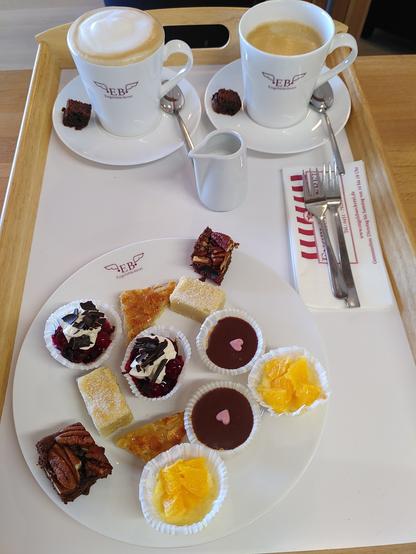 Auf einem großen Tablett steht ein Teller mit Kuchen im Miniaturformat (alle Küchlein sind je zweimal darauf): Schokotörtchen mit Earl-Grey-Tee, Orange Cheesecakes, Florentiner, Schwarzwälder-Kirsch-Brownies, Vanilla Shortbread und Brownies mit Pekannüssen. 
Dahinter stehen zwei Becher Kaffee auf Untertassen. Davor steht ein Kännchen mit frischer Milch.