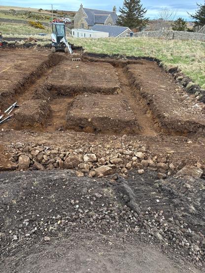 A photo of an excavated area. The sandy earth has a series of trenches dug into it that vaguely show the layout of a small house. A small digger is sitting at the end of the area with grassy fields and neighbouring buildings beyond.