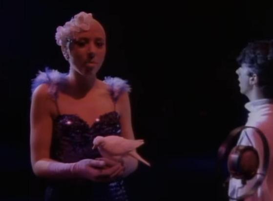 A bald woman in a sequinned dress with flowers on her head sings into a very old microphone, she has a dove in her hands. Behind Marc Almond wearing a white top also sings facing away from her. A screen shot from the video for Sofy Cell’s Torch single