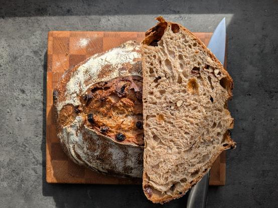 Bread loaf, laying on a wooden cutting board with checker pattern, laying on grey kitchen desk. The loaf is cut in half, one half has the cut positioned towards cam. Under this half a silver kitchen knife resides. Sun is coming from the top of the snap.