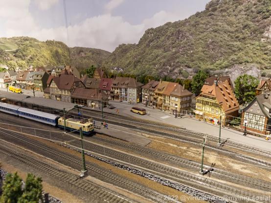 A photo of a model railway. A trainstation with an old electric passenger train can be seen. Behind the train station is a small village with some olf half-timbered hoses and old busses.