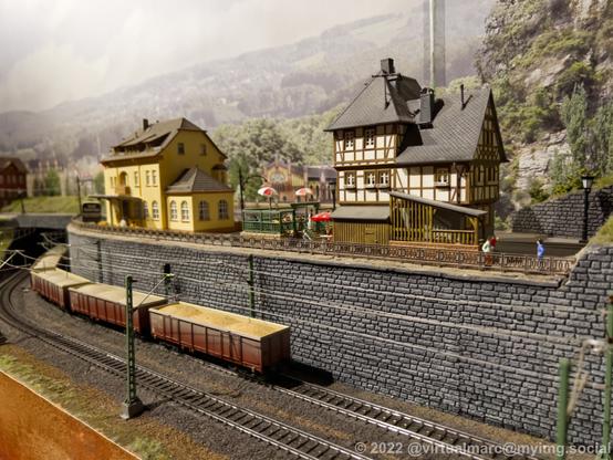 A photo of a modelrailway. A fright train with loaded sand is passing by. Behind are some old houses, looking like restaurants.