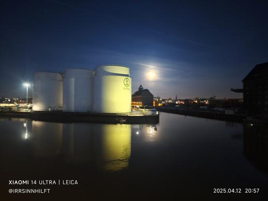 3 Silos oder Tanks auf einer Insel im Westhafen spielgeln sich durch Laternen und dem hellen Mond im glatten Wasser des Hafens. Dahinter eine Skyline aus Hafen-Speicher und der Stadt