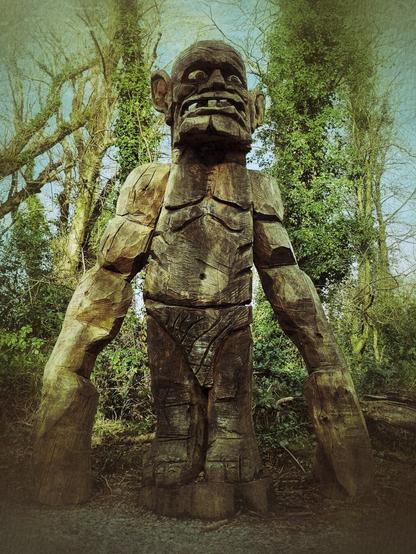 A large, weathered wooden sculpture of a troll with exaggerated features, including very long arms, stands in a forest clearing.