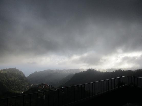 Another early morning, looking at the ridges of Santana (right through the middle in the back) and São Jorge (that bump on the left) and the valley below, covered in patches of sunshine coming from below the clouds, with fluff of rain glowing in the light. To the right ,still in the dark, the small wood is steaming, while the whole sky is full of dark grey clouds hanging low, like a heavy blanket covering the island.