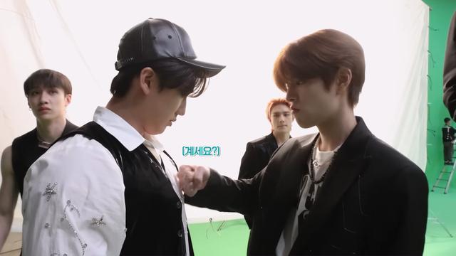 Seungmin raises a fist to knock it against Changbin's chest. Changbin's looking down at Seungmin's fist. Bang Chan is walking away and not looking at either of them. He has a flat expression. Hyunjin's in the background, biting his lip as he looks at Changbin's chest.