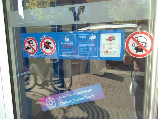 Nahaufnahne einer angeschnittenen  Glastür im Querformat. Oben ein Milchglasstreifen mit ausgeschnittenem Volksbank-V. Darunter ein breiter blauer Aufkleber, darauf von links nach rechts:
1. Rotes Verbotsschild mit durchgestrichener Fotokamera, Videokamera und Smartphone mit aktiver Kamera.
2. Rotes Verbotsschild mit durchgestrichenem Motorradhelm.
3. Infokasten „Geldbestände Zeitschloss-gesichert“ mit etwas Zusatztext.
4. Infokasten mit Überwachungskamera-Symbol und Kleingedrucktem.
5. Ein optisch abweichender Infokasten mit kleingedrucktem Text  in 5 Sprachen mit der Info „Autonome Gasneutralisierung“.

Rechts neben dem blauen Aufkleber ein separates rotes Verbotsschild mit durchgestrichenem Kopf mit Mund-Nase-Schutz.

Weiter unten schräg aufgeklebt ein großer Hinweis: „Follow us on Instagram: deine_heimatbank“.