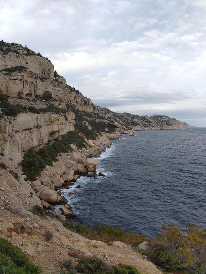 Après le pas du pêcheur, vue plein Est au dessus de la calanque de l'Escu (encore une que je n'avais jamais visité jusqu'à présent).

Sur la gauche on peut voir des falaises de calcaire, entrecoupés par de vires avec de la végétation assez dense. Au pied de ces grands murs on voit des énormes blocs de pierres détachés qui ont atteint le rivage de la mer.

Sur la droite l'étendue d'eau d'un bleu foncé (car le ciel est couvert).

En fond, et en continuaison avec cette côte de falaise, on peut voir toute la pointe de Sormiou (côté sud-ouest) non visible par les habituée de la calanque du même nom.