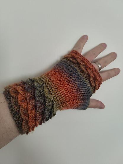 A hand wearing a fingerless crochet mitten featuring a dragon-scale pattern. The yarn transitions through shades of orange, red, green, and purple, creating a vibrant yet slightly muted effect. The glove extends to cover part of the forearm, with textured layers resembling overlapping scales. A silver ring is visible on the ring finger, and the background is a neutral, light-colored surface.
