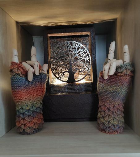 Two wooden mannequin hands are displayed within a wooden shelf, each adorned with fingerless crochet gloves featuring a dragon-scale pattern. The yarn shifts through shades of orange, red, green, and purple, creating a textured, mythical effect. Between them, a small illuminated fountain with an intricate tree design casts a warm glow, adding to the cozy, artistic ambiance.