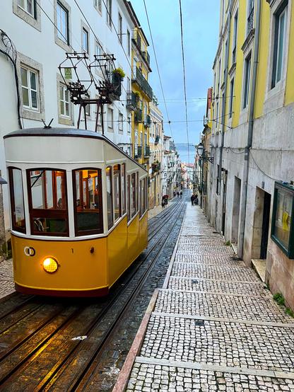 A yellow vintage tram is traveling down a narrow, cobblestone street lined with classic Portuguese buildings. The scene is set on a rainy day, with a cloudy sky and a view down the street towards the horizon. There are a few people walking along