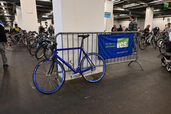 Rennvelo mit blauem Rahmen und blauen Felgen vor einer VCS-Fahne mit blauem Hintergrund.
