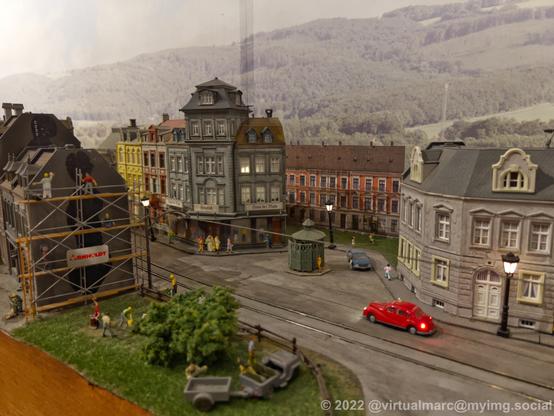 A photo of an old city of a model railway with old cars in the streets. There is a tram line and some shops. One house is currently renovated.
