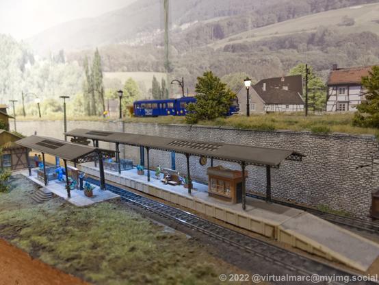 A photo of a small model railway train station, wirh a small kiosk on one platform. A bus is driving on a road behind the station.