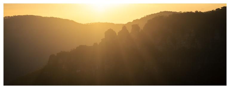 A wide-format color photo with a white border, capturing a mountainous landscape bathed in warm, golden sunlight. Layers of hills stretch across the frame, silhouetted softly by the backlighting sun. Prominent rock formations stand out distinctly, surrounded by sunbeams and glowing haze.