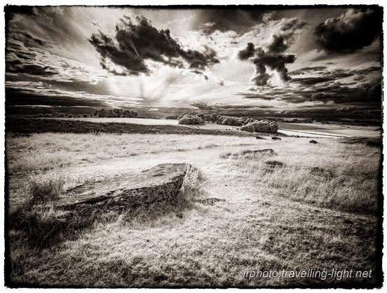 A toned black and white infrared photo of stone slabs lying in a grassy area of moorland, against a dramatic sky of sunlight and broken cloud.