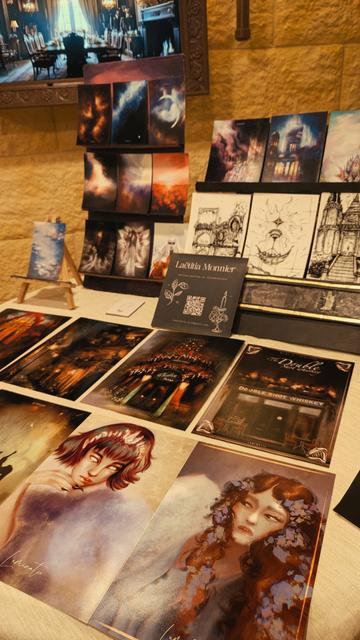 Photo d'un stand d'illustration au marché de créateurs organisé par le Malleus Malificarum. On y voit des illustrations fantastiques, d'architectures oniriques et des portraits de jeunes femmes aux allures d'elfes.