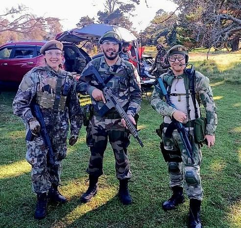 Tres personas, dos chicos y una chica, posando para la foto con equipamiento militar completo. Al fondo se pueden ver coches aparcados y más jugadores de airsoft preparándose para una partida.