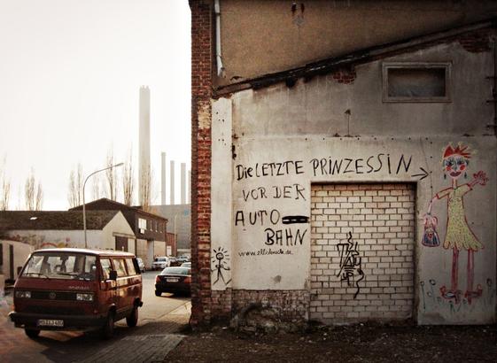 A photo in warm retro-brown colours shows a run-down industrial area. An old red VW Bulli van is parked at the side of the road, "the last princess before the motorway" is painted on the wall of a dilapidated building, next to it a childish-looking drawing of a woman with a crown.