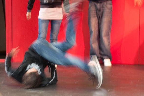 A hip hop girl in a sort of somersault on the ground, 1 jeans leg in the air, the other seems to follow. The image of the girl is blurred by the movement. In the background the legs of 3 other hip hoppers. they are standing against a red back wall.