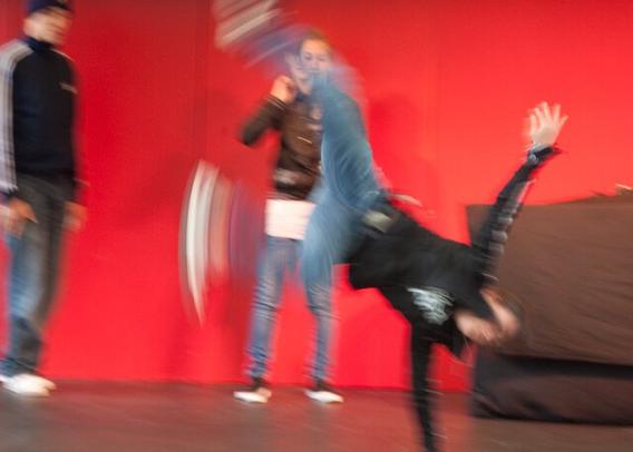 The same girl but now standing on one hand, the other arm in the air. The movement makes the legs blurry. In the background her two friends in jeans against a red background.