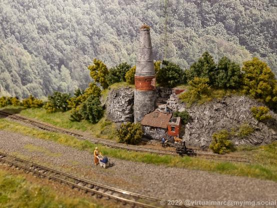 A photo of a model railway. Between two rails a man is walking along with a stroller. Behind the railway is an old brick building with a large chimney which houses a nest with storks.