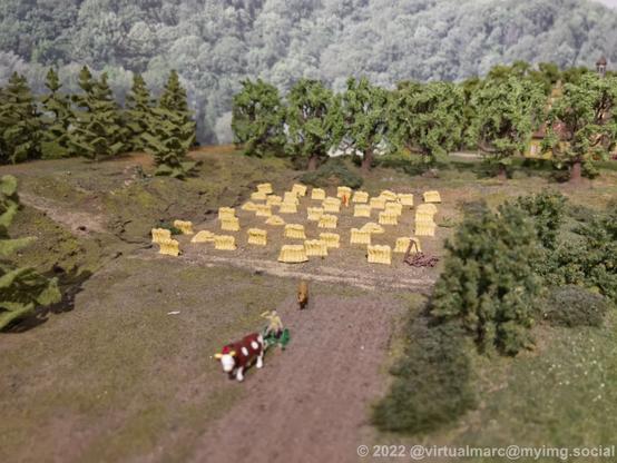 A photo of a model railway. A farm can be seen with weed. A farmer is working with a cow on the farmland.