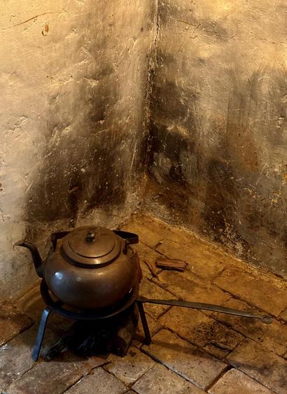 A brown-toned photo of an antique copper kettle in a fireplace (at Ephrata Cloister, Pennsylvania). The kettle sits at the lower left of the image, on a trivet with a long handle that stands over some kindling on a brick floor. The walls of the fireplace bear dark scorch marks from past fires.