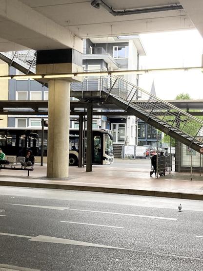 Zu sehen ist ein Ausschnitt vom Siegburger Busbahnhof. Von links fährt ein schwarz weißer Bus hinter einer Betonsäule los. Im Vordergrund die Fahrbahnen im Inneren des Busbahnhofs. Rechts im Bild führt eine Treppe nach links oben. Im Hintergrund ein Haus und ein kleines Spitzchen Kirchturm.