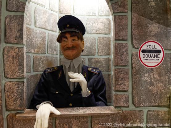 A photo of a conductor statue. It's hands and head were moving. A customs sign for the border to italy is hanging next to it.