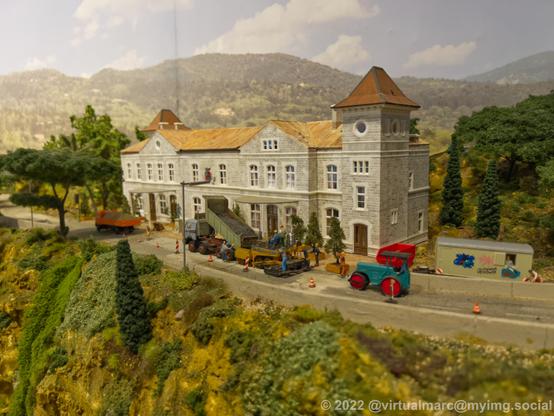 A photo of a big brick building on a model railway. It's standing next to a street with trucks. There is a roadwork going on.