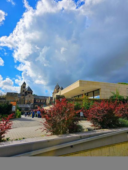 Blick auf eine moderne Architektur im Vordergrund mit einer historischen Kirche im Hintergrund, umgeben von grünen Pflanzen und einem bewölkten Himmel.