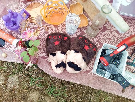 A blanket on the ground with used items laid out for sale. There's a vase, some shoe spreaders, a kind of juicer, long expired fire resistant spray foam, a tub of old TV remotes and in the center two big slippers that look like faces wearing a dark fur hat. They have thick black eyebrows and a mustache. The hat has a red star on it and SSSR written in cyrillic.