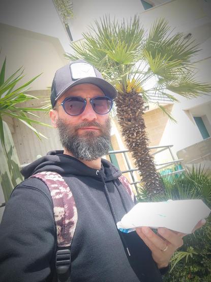 @adrianobono Un uomo con barba e occhiali da sole scuri indossa un cappellino nero con una patch bianca e un cappotto nero. Porta una borsa a tracolla con una tracolla a motivi marroni e bianchi. Sta tenendo un oggetto bianco, probabilmente un pacchetto, nella mano destra. Sullo sfondo, si vede un palma e un edificio bianco con una ringhiera metallica. L'atmosfera sembra essere di un giorno luminoso e soleggiato.
―