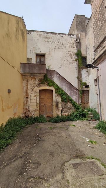 L'immagine mostra un cortile interno di un edificio antico, caratterizzato da pareti invecchiate e deteriorate. A sinistra, una parete gialla con segni di usura e una finestra con tende chiuse. Al centro, un portone di legno scuro con una cornice in pietra, con un balcone sopra. A destra, una scala in cemento con rampa e piante rampicanti che arrampicano lungo le pareti. Il pavimento è di terra battuta con erbacce e detriti. Un lampione è visibile in alto a destra.
―