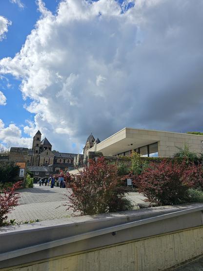 Das Bild zeigt eine malerische Szene mit einem historischen Gebäude im Hintergrund, das von einer modernen Struktur flankiert wird. Der Himmel ist teilweise bewölkt, mit großen, weißen Wolken, die einen kontrastierenden Hintergrund bilden. Im Vordergrund sind gepflegte Sträucher mit roten Blättern zu sehen, die das Bild mit Farbe bereichern. Ein gepflasterter Weg führt zu einem Gebäudekomplex, das eine Mischung aus gotischen und modernen architektonischen Elementen aufweist. Einige Menschen sind auf dem Weg zu oder von dem Gebäude, was auf eine touristische oder öffentliche Nutzung hinweist. Die Kombination aus historischer und moderner Architektur sowie die natürliche Umgebung vermittelt ein Gefühl von Ruhe und Kultur.
Bereitgestellt von @altbot, privat und lokal generiert mit Ovis2-8B
