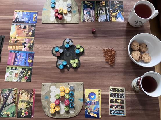 Zu sehen ist ein dunkel brauner Tisch auf dem das Brettspiel Harmonies gespielt wird. Vom Spiel liegen fünf Tierkarten aus und zwei Spielbretter der Spielenden. In der Tischmitte sind verschiedenfarbige Spielsteine. Am rechten Rand stehen zwei Tassen mit Tee und dazwischen eine Schale mit Keksen.